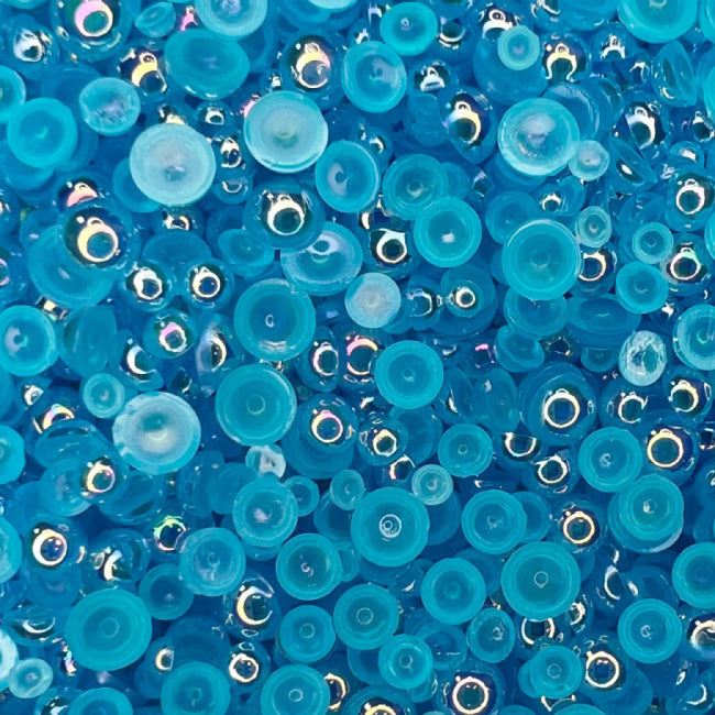 A close-up view of blue transparent iridescent acrylic pearls intended for crafting and embellishment purposes, with the text 'RAZZLEBERRY BUBBLES Transparent Iridescent Baubles Embellishment Mix' and the website 'www.trinitystamps.